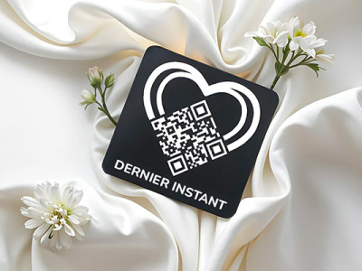 Médaille funéraire Dernier Instant avec QR code, posée sur un tissu blanc et des fleurs, pour préserver la mémoire d’un proche décédé